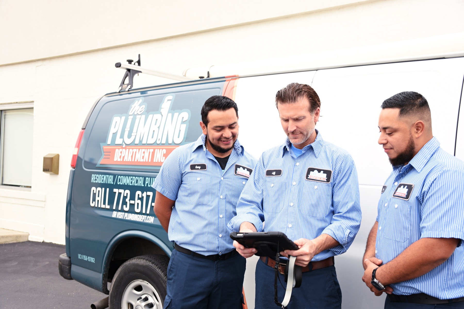 A Plumbing Department lead plumber reviewing a job on a tablet with two technicians beside a company van