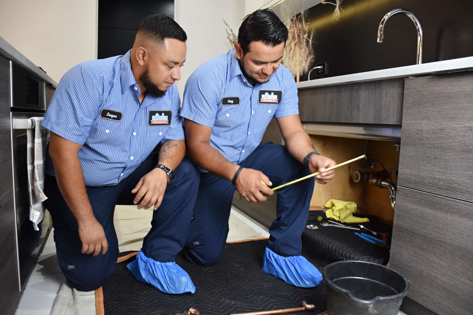 A senior plumber kneeling beside a newer plumber, pointing out a detail under a kitchen sink